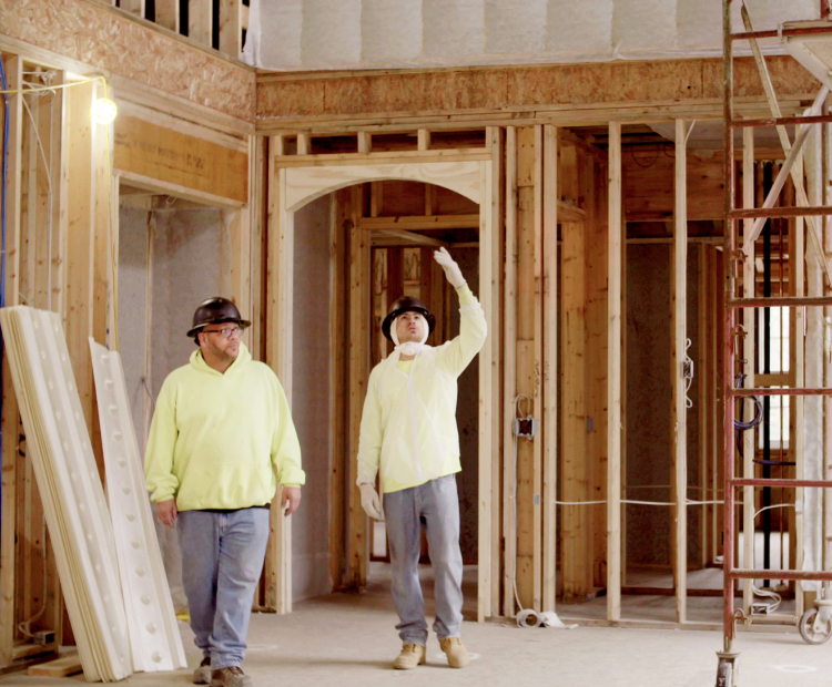 Two contractors in pale yellow sweatshirts, jeans, and hard hats, standing in a mid-construction home, one of them looking and pointing upward.